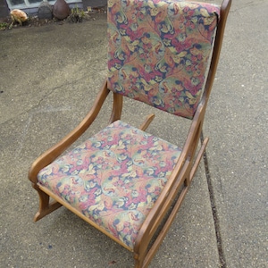 May include: A wooden rocking chair with a floral patterned fabric seat and back. The chair has a curved back and a wide, comfortable seat.