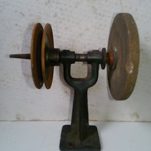May include: An antique grinding wheel with a dark green metal frame and a stone grinding wheel. The wheel has a wooden pulley on one side and a stone grinding wheel on the other. The base is square.