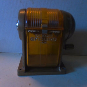 May include: A vintage Dixon Enduro pencil sharpener. The sharpener has a brown base and a transparent amber-colored top. The text "PUSH" and "DIXON ENDURO" are visible on the front. A small silver handle is on the side.