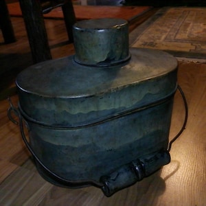 May include: An antique metal container with a cylindrical top and a large, oval-shaped body. The container has a dark, aged patina with a handle and a cylindrical handle on the side. The container is likely used for storage or carrying.