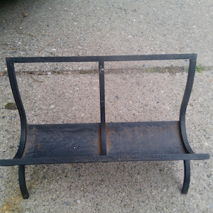 May include: Black metal fireplace log holder. The holder has a rectangular base with a vertical divider and curved sides. The overall design is simple and functional, suitable for storing firewood near a fireplace.