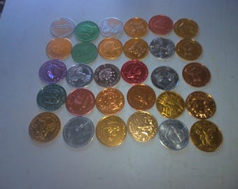Könnte beinhalten: Eine Sammlung von 25 runden, metallischen Spielmarken mit verschiedenen Designs und Farben. Die Spielmarken sind in einem Raster auf einer weißen Oberfläche angeordnet.