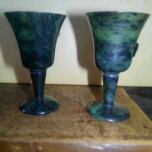May include: Two green and black marble goblets with a flared top, slender stem, and round base. The goblets are displayed on a wooden surface. The marble has a natural, mottled pattern.