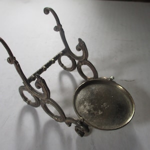 Op de afbeelding: Een zilverkleurige metalen standaard met een sierlijk ontwerp. De standaard heeft een ronde bovenkant en drie poten. De standaard is gemaakt van metaal en heeft een zilveren afwerking.