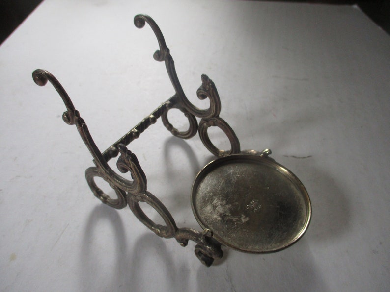 Op de afbeelding: Een zilverkleurige metalen standaard met een sierlijk ontwerp. De standaard heeft een ronde bovenkant en drie poten. De standaard is gemaakt van metaal en heeft een zilveren afwerking.
