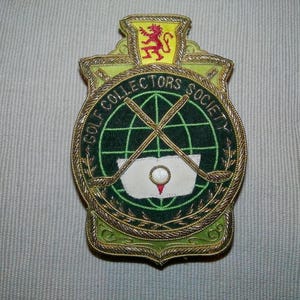 May include: Embroidered patch with a shield shape, featuring the text "GOLF COLLECTORS SOCIETY" around a green globe with crossed golf clubs. The patch has gold trim and a crest with a red lion.