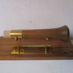 May include: A wooden secretary caller with brass accents, mounted on a wooden base. The caller has a flared end and a mouthpiece. The base has a brass plate with the text "SECRETARY CALLER DON'T YELL BLOW".