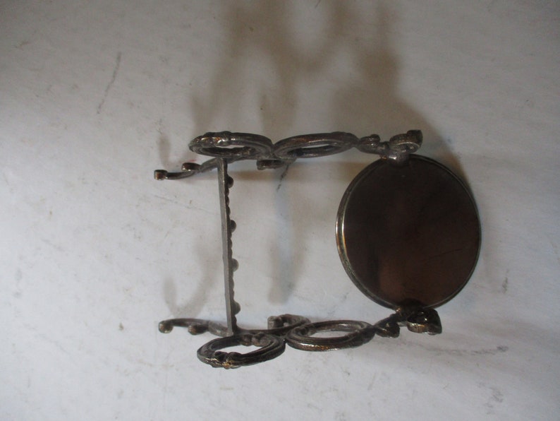 Op de afbeelding: Een decoratieve standaard van zilverkleurig metaal met een ronde bovenkant en drie poten. De standaard heeft een decoratief ontwerp met krulletjes en lussen.