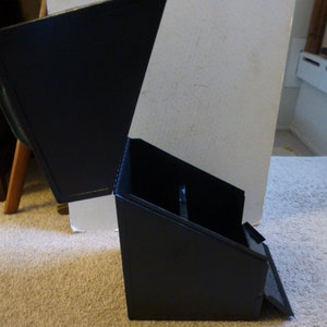 May include: A black, angled, cardboard box with a hinged lid. The lid is open and the box is sitting on a beige carpet.