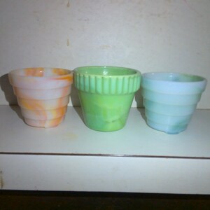 May include: Three small, decorative flower pots in shades of orange and white, green, and blue. The pots have a tiered design. The orange and white pot has a marbled effect. The green pot has vertical ridges. The blue pot has a swirl effect.