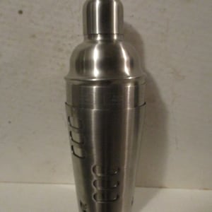 K&ouml;nnte beinhalten: Ein Cocktailshaker aus Edelstahl mit abgerundeter Oberseite und zylindrischem Boden. Der Shaker hat eine Reihe von Aussparungen und ist zum Mixen von Getr&auml;nken konzipiert. Die Metalloberfl&auml;che reflektiert das Licht.