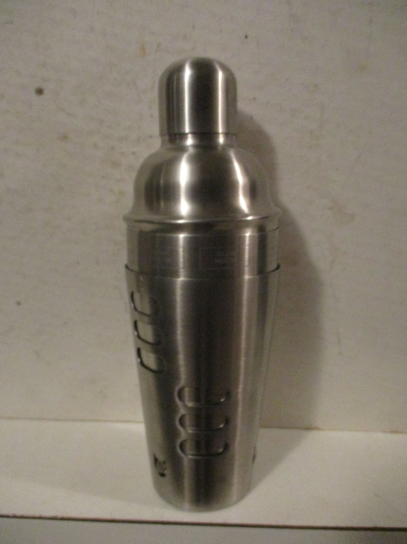 K&ouml;nnte beinhalten: Ein Cocktailshaker aus Edelstahl mit abgerundeter Oberseite und zylindrischem Boden. Der Shaker hat eine Reihe von Aussparungen und ist zum Mixen von Getr&auml;nken konzipiert. Die Metalloberfl&auml;che reflektiert das Licht.
