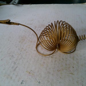 May include: A gold-colored metal alligator clip with a coiled spring design. The clip is attached to a curved arm that leads to a heart-shaped coil. The spring is approximately 10 cm long.