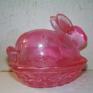 May include: A pink glass bunny-shaped container with a basket-weave base. The bunny has upright ears and a rounded body, and the container is likely for Easter or spring decor.