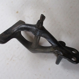 May include: A black metal clamp with a spring and a screw. The clamp is designed to hold two pieces of wood together.