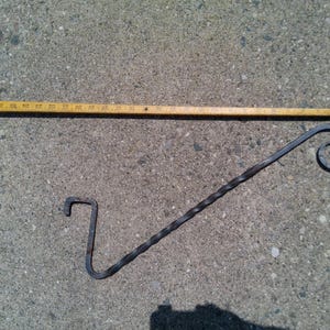 May include: A hand-forged, dark metal plant hanger with a twisted stem and a curled end. The hanger is approximately 12 inches long. A wooden ruler is also in the image.