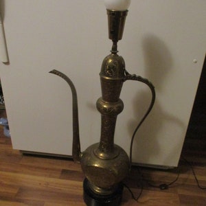 May include: A vintage brass lamp with an ornate, jug-like design. The lamp features a long spout, a handle, and a bulb at the top. It sits on a black base and has a power cord. The lamp's surface is detailed with etched patterns.