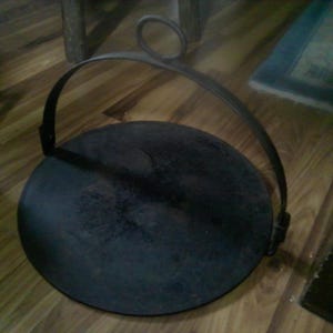 May include: A round, flat, black metal griddle featuring a curved handle that arches over the top. The handle includes a circular loop at the apex for hanging. The surface of the griddle is dark, textured, and shows signs of use and wear.