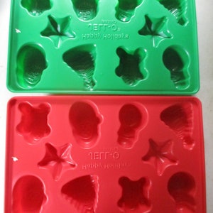 May include: Two plastic Jell-O brand molds for making holiday-themed gelatin desserts. The top mold is green and the bottom mold is red. Both molds have a variety of shapes, including stars, trees, and ornaments. The molds are labeled "Jell-O Brand Happy Holidays".