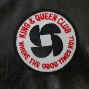 May include: A white and red embroidered patch with the text "King & Queen Club Where The Good Times Roll" in red lettering and a black stylized flower design in the center.