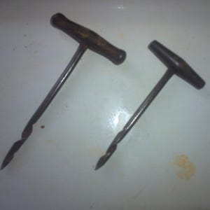 May include: Two antique hand-crank drills with wooden handles. The drills have a metal spiral bit and a metal T-shaped handle.