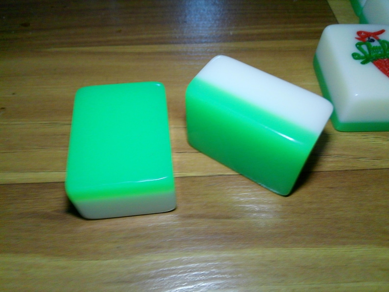 Pu&ograve; includere: Due tessere rettangolari da Mahjong con una finitura liscia e lucida. Una tessera &egrave; posizionata verticalmente mostrando una parte superiore verde brillante e una base bianca, mentre l'altra &egrave; inclinata per rivelare la parte superiore bianca e la base verde. &Egrave; visibile una terza tessera con il disegno di un uccello rosso.
