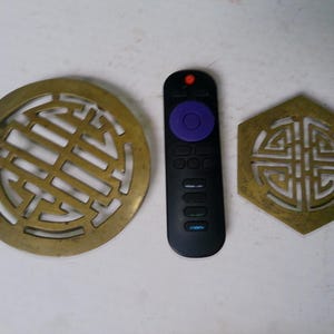 May include: A circular brass trivet with an openwork geometric pattern, a black remote control with a purple circular navigation button and a red power button, and a hexagonal brass trivet featuring a matching openwork geometric design.
