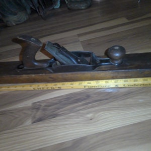 May include: A vintage wooden hand plane with a dark finish, featuring a handle and a knob. The metal blade is visible. A yellow ruler with black text is below the plane. The text on the ruler reads "CHECK'S Furniture & Undertaking."