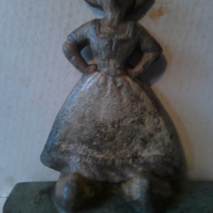 May include: A vintage cast iron doorstop in the shape of a woman wearing a white dress and a hat. The doorstop is painted in shades of gray and white and has a green base.