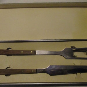 May include: A set of two kitchen utensils with wooden handles. The first utensil is a carving fork with a metal handle and a wooden handle. The second utensil is a carving knife with a metal handle and a wooden handle. The utensils are in a white box.