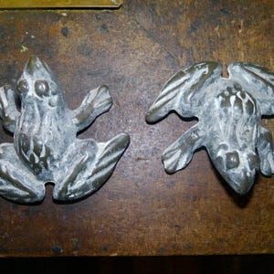 May include: Two antique bronze frog-shaped door knockers. The frogs are detailed with textured skin and outstretched arms and legs. The door knockers are mounted on a wooden surface.