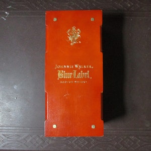Pu&ograve; includere: Una scatola rettangolare in legno con una vivace finitura arancione. La scatola presenta le parole "JOHNNIE WALKER, Blue Label, scotch whisky" in lettere dorate. Un emblema dorato &egrave; in alto. La scatola ha accenti angolari color ottone.