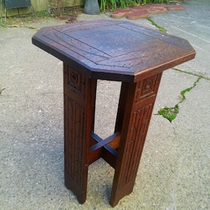 May include: A dark brown wooden side table with an octagonal top featuring carved geometric patterns. The table has four vertical legs with carved square motifs at the top and a cross-shaped support brace connecting the legs near the base.