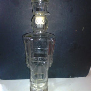 May include: A clear glass bottle shaped like a toy soldier. The bottle has a cork stopper and a hat on top. The bottle is empty.