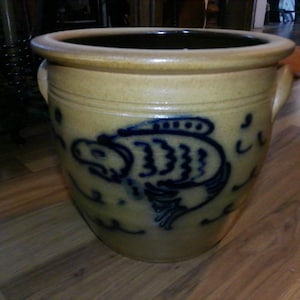 May include: A tan stoneware crock with a dark blue fish design. The crock has a rounded shape with two small handles and a dark interior. The fish is stylized with detailed fins and scales. A decorative piece for the home.