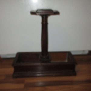May include: A dark brown wooden stand with a rectangular base and a central vertical post. The post supports a small, cross-shaped top. The stand appears to be for displaying an item. The wood has a polished finish.