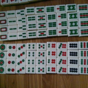 Pu&ograve; includere: Una collezione di tessere rettangolari bianche da Mahjong disposte in due file orizzontali. Le tessere presentano simboli, caratteri e motivi circolari incisi con inchiostro verde, rosso e nero. Le tessere hanno una finitura liscia e lucida.