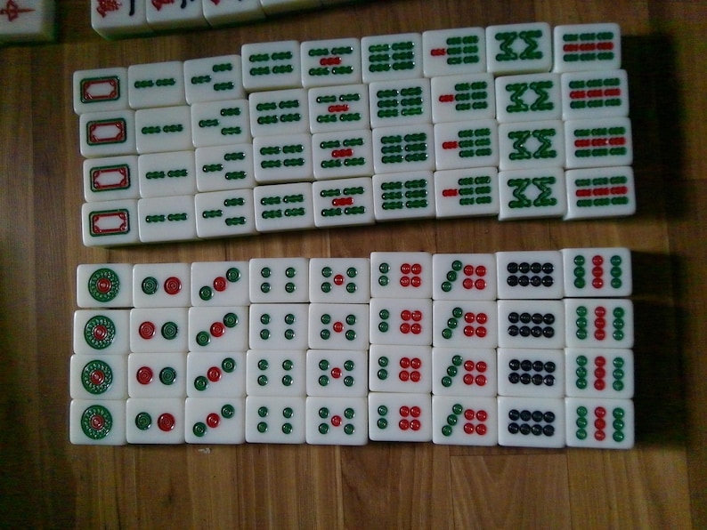 Pu&ograve; includere: Una collezione di tessere rettangolari bianche da Mahjong disposte in due file orizzontali. Le tessere presentano simboli, caratteri e motivi circolari incisi con inchiostro verde, rosso e nero. Le tessere hanno una finitura liscia e lucida.