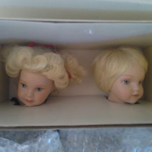 May include: Two porcelain doll heads with blonde hair. The girl doll has a red bow in her hair. The boy doll has short hair. Both dolls have closed eyes.