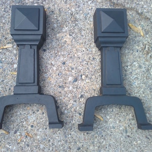 May include: Two black metal fireplace andirons with a square top and a pyramid-shaped design. The andirons have a decorative base with three legs.