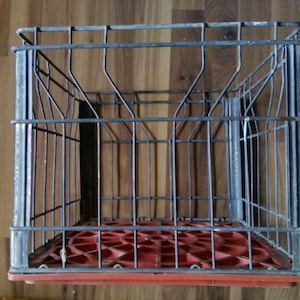 May include: A rectangular storage crate featuring a grey metal wire frame structure and a solid red plastic base. The wire frame consists of vertical and horizontal bars forming an open grid design, while the base has a circular pattern.