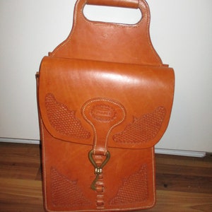 May include: A tan leather bag with a handle and a flap closure. The bag features decorative stitching and embossed patterns. A metal clasp secures the flap. The bag is designed to be carried by hand.