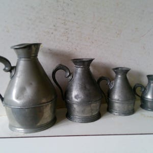 May include: Four antique pewter pitchers of varying sizes. The pitchers have curved handles and a dark, metallic gray color. The largest pitcher is on the left, with the smallest on the right. The pitchers are arranged in a row.