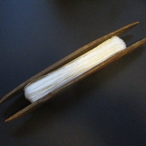May include: A wooden yarn swift, a tool for winding yarn into a ball. The swift is made of dark wood and holds a bundle of white yarn. The yarn is tightly wound and ready for use. The background is a dark surface.