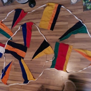 May include: A collection of colorful nautical signal flags, each with a unique design and color scheme. The flags are connected by a white rope, and are arranged on a wooden surface. The flags feature red, blue, yellow, green, and black.