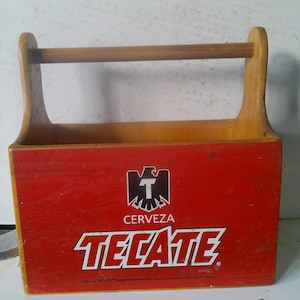 May include: Wooden beer caddy with a red front panel featuring the "Tecate" logo and the words "Cerveza Tecate". The caddy has a wooden handle and is designed to hold bottles.