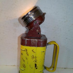 May include: A vintage rectangular lantern with a bright yellow body and dark red accents. It features a pivoting chrome-rimmed light head that is currently illuminated, a sturdy yellow handle on the side, and visible signs of wear on the metal finish.