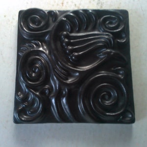May include: A black square tile with a raised design of swirling patterns and lines. The tile has a smooth, glossy finish.