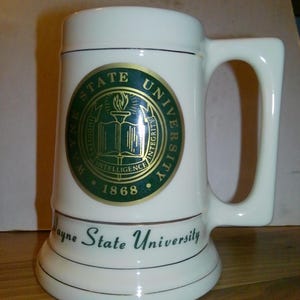 May include: A white ceramic beer mug featuring the Wayne State University seal in green and gold, with the text Industry, Intelligence, Integrity, and 1868. The words Wayne State University are printed in green script below the circular university seal.