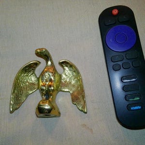 May include: A gold-colored eagle figurine with outstretched wings. The eagle is positioned next to a black remote control with a blue circular button and multiple smaller buttons.